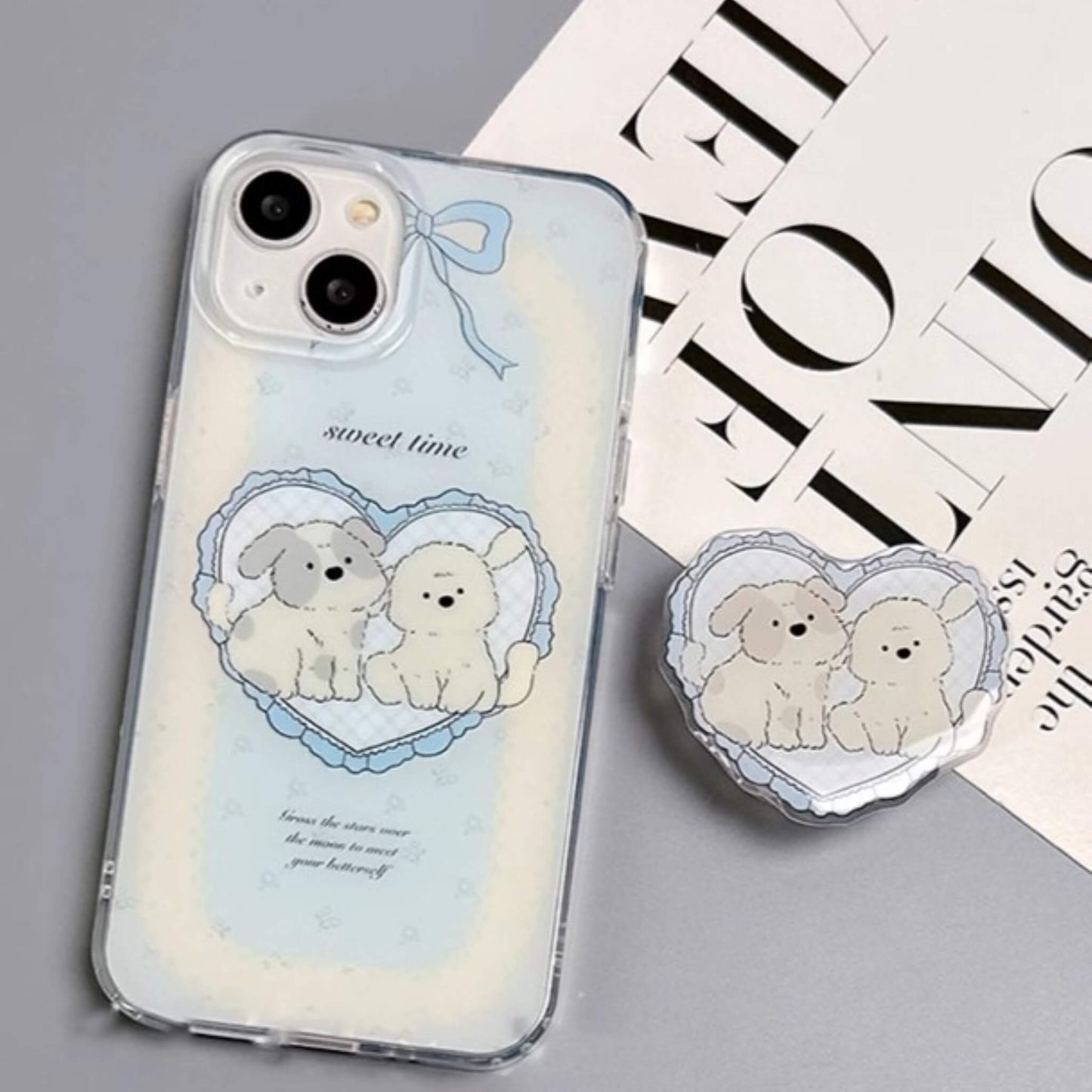 Charming MagSafe-compatible phone case displaying a heart with two cute puppies and 'sweet time' text, against a sky-blue background with soft clouds, next to a heart-shaped MagSafe popsocket with a matching design.