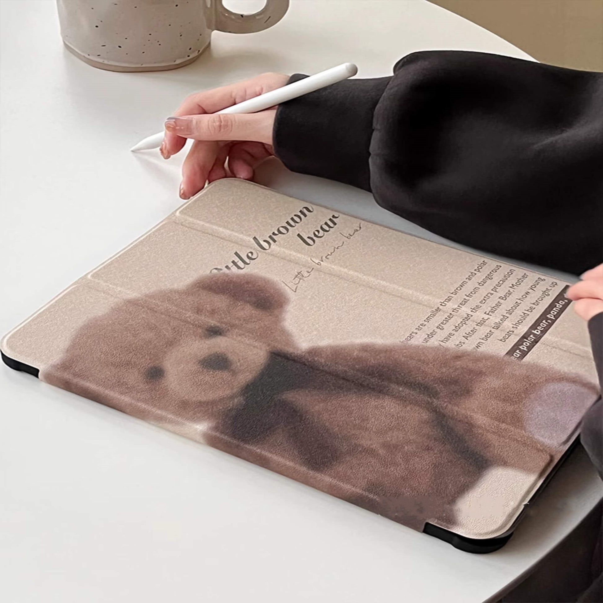 "Beige iPad case with a vintage teddy bear design and scripted text, displayed next to a white ceramic mug. Acrylic clear back with a slot for Apple Pencil."